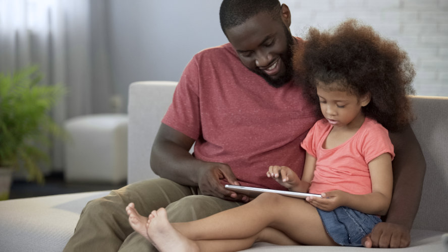 A father sits on a couch next to his daughter who is holding a tablet